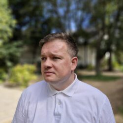  A person wearing a white collared shirt stands outdoors with trees and greenery in the background.